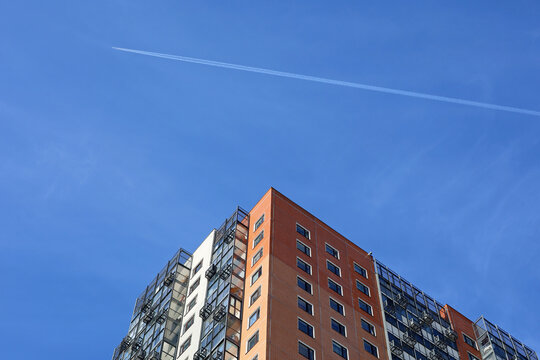 Jet airplane with white contrail flies over the residential building. Trace of the high flying plane on the blue sky - Powered by Adobe