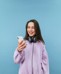 Positive teen girl in headphones around her neck and casual clothes enjoys smartphone and laughs, isolated on blue background.