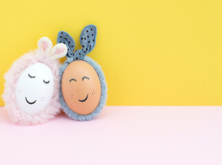 Two creative white and brown Easter eggs with bunny ears and painted eyes and mouth on a yellow and pink background.