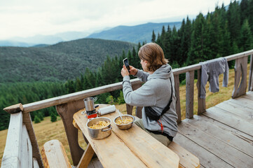 Male tourist sitting on the balcony of a bungalow in the mountains with food on the table and takes photos of beautiful landscapes on a smartphone camera.