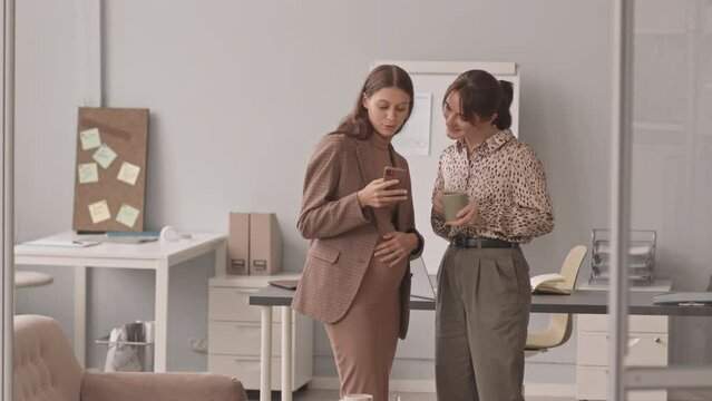 Medium slowmo shot of young Caucasian pregnant woman showing baby ultrasound image on smartphone to her colleague, standing at workplace in office during coffee break