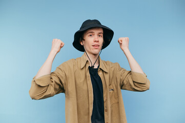 Portrait of a joyful boy in panama on a blue background with a smile on his face stands on a blue background and raises his hands up with joy.