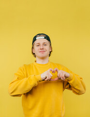 Portrait of a cute joyful guy in a sweatshirt and cap on a yellow background shows a gesture of love and looks at the camera and laughs.