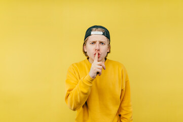 Funny guy in a cap shows a gesture of silence by touching his finger to his lips and looking at the camera on a yellow background.