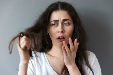 A young caucasian frustrated woman looks at the tips of her long dark hair. Gray background. The...