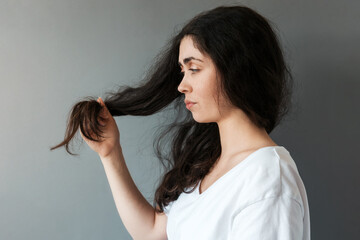 Fototapeta premium Portrait of young caucasian sad woman looks at the tips of her long dark hair. Gray background. Side view. The concept of hair care