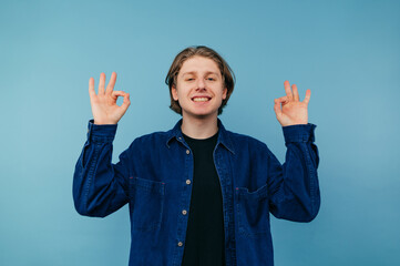 Positive guy in a shirt stands on a blue background with a smile on his face and shows his fingers...