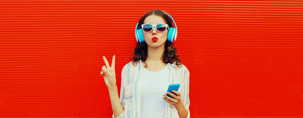 Portrait of stylish young woman posing in headphones listening to music with smartphone on red background