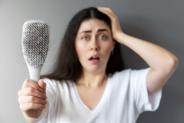 Blurred portrait of a shocked young woman shows her hairbrush with losing hair. Gray background....