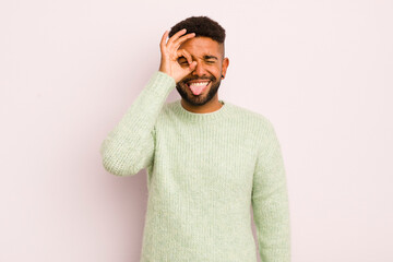 young afro man smiling happily with funny face, joking and looking through peephole, spying on...