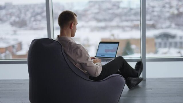 Busy male freelancer sits in bean bag chair with his back to camera. Man works on a laptop with graphs at the screen. Window with blurred cityscape at backdrop.