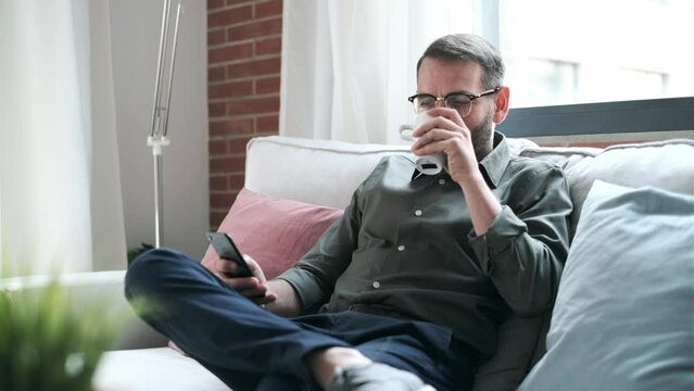 Video of mature male using smartphone texting while drinking a cup of coffee and relaxing on sofa at home.