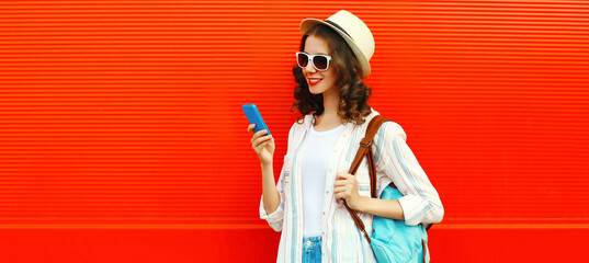 Portrait of happy smiling young woman holding phone wearing summer straw hat, backpack on red...