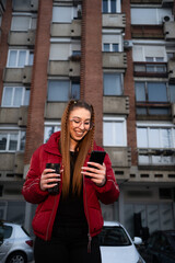 a young girl with glasses, in a red jacket, looks at the phone while holding a cup of coffee in her hand, in front of buildings in winter