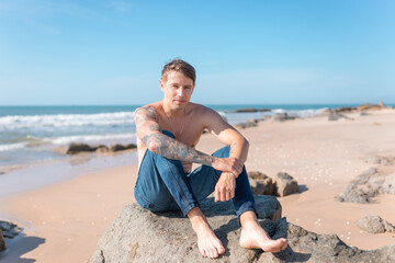 Attractive young caucasian bare-chested man sitting on the rock on the beach.