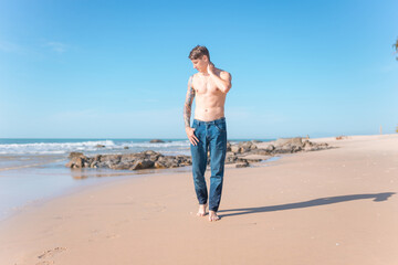 Attractive young caucasian bare-chested man walking on the beach barefoot.
