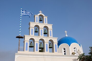Amazing white and blue greek orthodox church with religious crosses, bells and the greek national flag waving in the air against a blue sky in Santorini