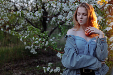 A young woman stands in an apple orchard and takes on an apple. Spring. Sunset. High quality photo