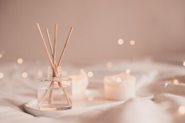 Home liquid fragrance in glass bottle and burning candles staying on white ceramic tray in bed...