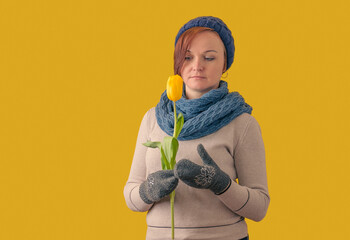 woman in a hat and mittens looks at a tulip flower on a yellow background