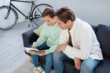 Smiling man sitting near son reading book on sofa in living room