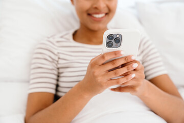 Young black woman smiling and using cellphone while sitting in bed