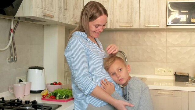 Pregnant mom with kid playing together and huging in the kitchen. Son listening to mom's pregnant belly