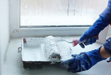 Close-up of hands in work gloves holding and collecting white paint in a bath. Paint the window slope with paint roller