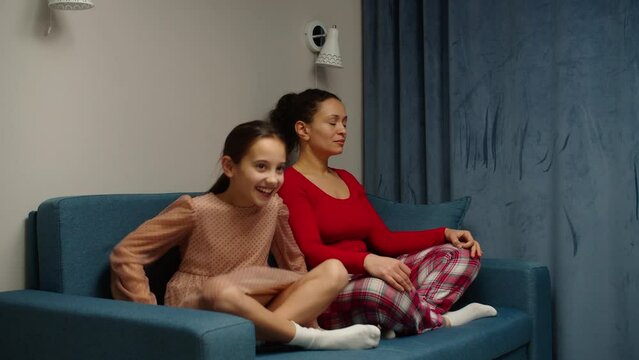 Mindful multiracial mom with cute funny daughter doing yoga exercise at home. Calm mother and preteen girl sitting in lotus pose on couch together. Mum teaching child to meditate. Moving shot