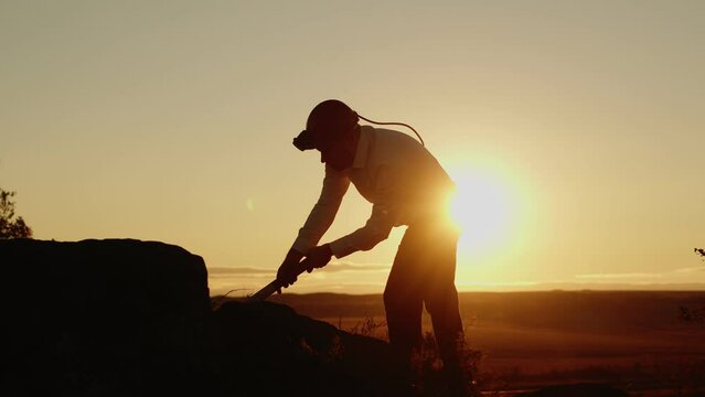 Silhouette man in helmet and white shirt hits stone with pickaxe background of sunset outdoor, side view. Businessman with pickaxe and helmet extracts ore from stone. Crypto Mining concept