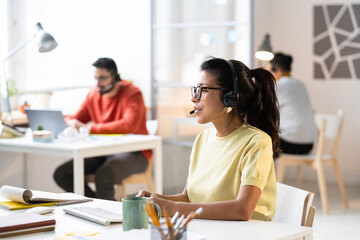 Young businesswoman in headphones with microphone working with customers and solving their problems while sitting at the table at office