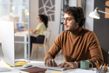 Call centre operator in headphones talking to the customer on the phone and typing on computer keyboard during his work at the table at office