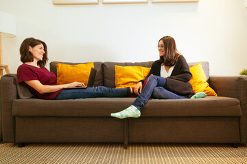 lesbian couple looking at each other and laughing on the couch with the laptop