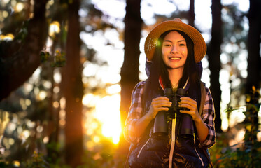 Asian young beautiful female tourist wearing hat, backpacking, holding binoculars, smiling with happiness for traveling at sunset in nature. Outdoor holiday, vacation and travel concept.