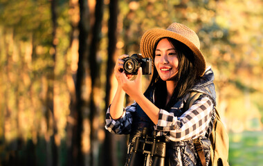 Asian young beautiful female tourist wearing hat, backpacking, holding camera, smiling with happiness for traveling at sunset in nature. Outdoor holiday, vacation and travel at the golden hour.