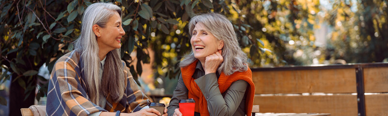 Mature Asian lady with positive grey haired friend spend time together sitting at small table in street cafe on nice autumn day