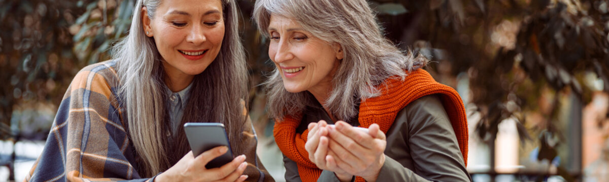 Asian Lady Shows Photos On Mobile Phone To Silver Haired Friend With Cup Of Tea Sitting At Small Table In Street Cafe