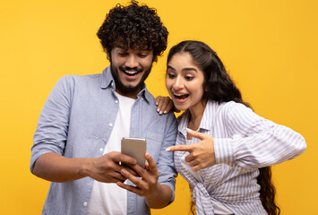 Excited indian couple using cellphone and pointing on it, looking at phone with open mouth, yellow background