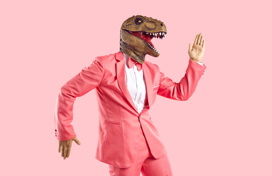 Funny man in rubber dinosaur mask dancing and having fun in the studio. Happy lizard headed guy in stylish funky vibrant pink party suit doing Egyptian dance moves isolated on pink colour background