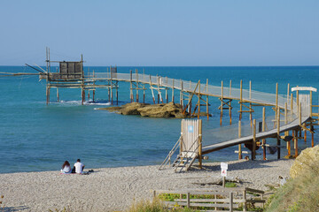 Punta Aderci Nature Reserve - Costa dei trabocchi - Abruzzo - A couple sitting on a pebble beach relaxes looking at the sea. In the background the imposing trabucco an ancient fishing machine.