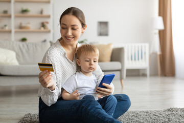 Millennial mother with baby boy using cellphone and credit card for online shopping, enjoying purchasing from internet