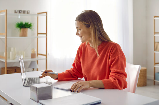 Young smart and successful woman working on laptop sitting at her workplace in office. Side view of smiling woman sitting at table with notebook and looking for information online or sending emails.