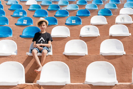 Serious boy in summer clothes sitting on one of chair on stadium and looking to camera.