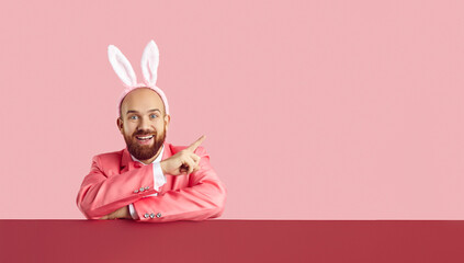 Look here. Funny happy cheerful excited bearded man in pink suit and Easter Bunny ears looking at camera and pointing to side at something on blank colour advertising copyspace copy space background