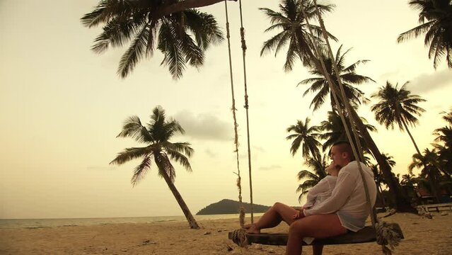 Cute loving couple on the swing on tropical beach. Woman and man in white shirt enjoy gold sunset and life in beaches, palm tree. Evening warm sunset. Summer holiday vacation tropical tourism concept