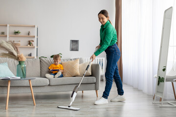 Fototapeta na wymiar Happy young mother mopping the floor and tidying up while her son sitting on sofa and watching cartoon on smartphone