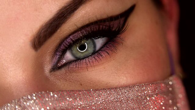 Woman in shimmer protective facial mask. Macro. Glitter eyeshadows and false lashes. Model eye blinking with fancy arrow make-up. Girl, green eye. Pandemic, covid-19 concept.