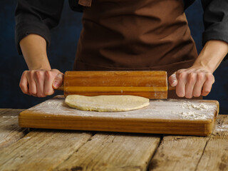 The chef rolls out the dough with a rolling pin on a wooden cutting board. Close-up. Minimalism. Wooden texture, dark background. Preparation of dough products. Advertising, banner.