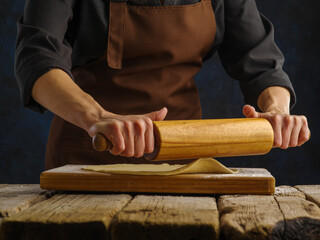 The chef rolls out the dough on a wooden cutting board with a rolling pin. Close-up. Recipes for confectionery, pizza, pasta, spaghetti. Home and restaurant cuisine, culinary blog.