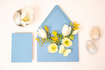Stationery flat lay with blue envelope and fresh spring tulip, mimosa nad ranunculus flowers